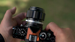 Hands holding an orange and black mirrorless camera with a telephoto lens mounted, shown outdoors against a blurred green background.