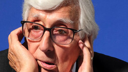 Older man with white hair and glasses against a blue background, hand near his face in a contemplative pose.