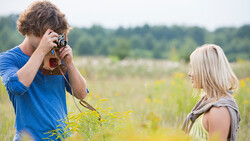 Young man with curly brown hair photographing a blonde woman in a sunlit field with vintage camera.