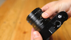 Hand holding a compact mirrorless camera with an attached prime lens against a warm wooden background.