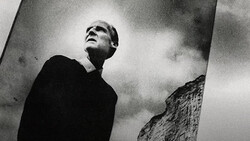 Man in dark clothing gazing upward against dramatic cloudy sky in high-contrast black and white photography.