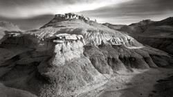 Dramatic black and white landscape of layered, eroded badlands with structures atop the highest ridge.