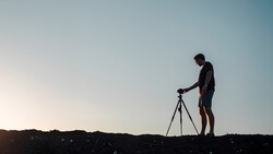 Silhouetted photographer operating a tripod-mounted camera against a clear sky at dusk.