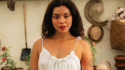 Woman with dark wavy hair and red lipstick wearing a white camisole in a studio setting.