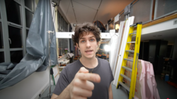 Person pointing at camera inside a narrow RV or camper van with storage shelves and equipment visible.