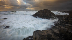 Waves crash over dark columnar basalt rocks jutting into turbulent sea at golden hour.