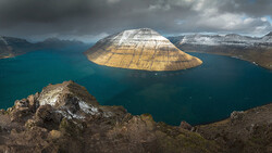 Snow-capped mountain with distinctive dome shape rising from turquoise fjord waters, viewed from rocky foreground.