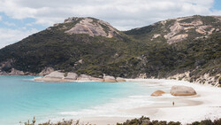 Turquoise bay with white sand beach, large boulders, and forested hillside under partly cloudy sky.