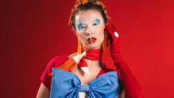 Portrait of a woman with blue eyeshadow and red lipstick against a red background, wearing a red jacket and blue bow.