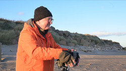 Photographer in orange jacket holding a camera on a beach with rocky cliffs in background.