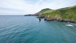 Rugged green coastline with steep cliffs meeting turquoise ocean water under partly cloudy sky.