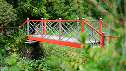 Bright red footbridge with X-pattern railings spanning a small stream in a lush garden.