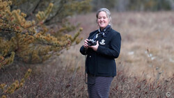 Female photographer holding a camera in an open field with sparse vegetation and evergreen shrubs.