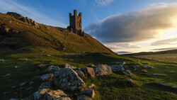 Medieval stone tower ruins on a hillside with golden hour sunlight and dramatic clouds.