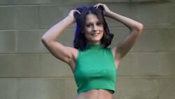 Woman in green crop top with dark blue hair against neutral brick wall background.