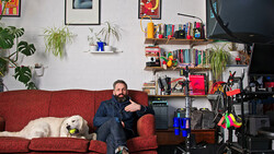 Man sitting on red couch with white dog in home studio setup with shelving and camera equipment.