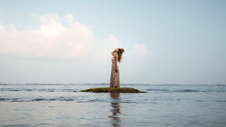 Woman in patterned dress standing on a wooden post jutting from calm shallow water under partly cloudy sky.