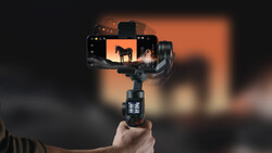 Hand holding a smartphone gimbal stabilizer with mounted phone displaying sunset silhouette footage.