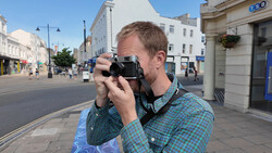 Man in checkered shirt holding vintage rangefinder camera to his face on a sunny urban street.