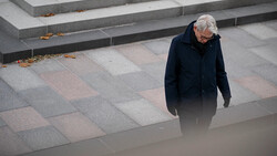 Elderly person in dark coat walking alone across geometric paved plaza with stepped levels.