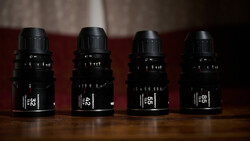 Four black cinema lenses arranged in a row on a wooden surface against a burgundy backdrop.