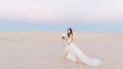 Bride in flowing white gown holding bouquet on sandy beach at dusk.