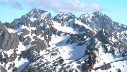 Snow-covered alpine peaks with forested slopes and exposed rock faces under partly cloudy skies.