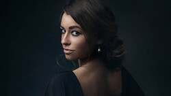 Woman with dark hair and hoop earrings looking over her shoulder against a dark background.