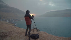 Photographer with tripod-mounted camera on hillside overlooking alpine lake at dusk.