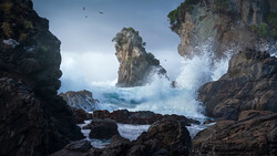Dramatic ocean waves crashing against rocky sea stacks and cliffs under a partly cloudy sky.
