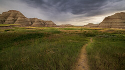 Dirt trail cutting through grassland with layered buttes under dramatic storm clouds.