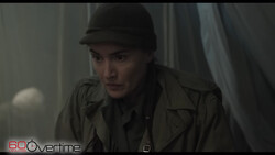 Person wearing dark fedora hat and olive jacket in dimly lit interior space.