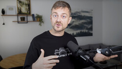 Man in black camera-themed t-shirt speaking expressively while holding a microphone in an indoor studio setting.