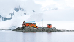 Bright orange research station building on a rocky outcrop in polar waters, with snow-covered mountains and glaciers in the background.