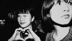 Two young women in a black and white portrait; one makes a heart shape with her hands while the other gazes downward.