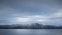 Misty mountain landscape across still water under overcast sky.