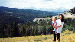 Woman holding a toddler standing in a mountain meadow with forested valleys and snow-capped peaks in the background.