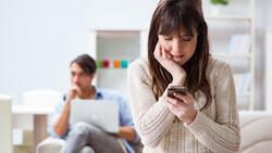 Woman in cream cardigan smiling while looking at smartphone, with man blurred in background working on laptop.