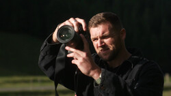 Photographer holding binoculars to his face against a dark background.