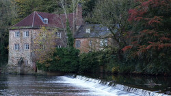 Historic stone mill building beside a river weir with autumn foliage in soft natural light.