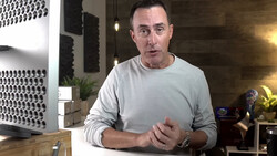 Man in light gray long-sleeve shirt speaking to camera in modern home studio setting.