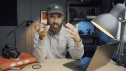 Bearded man in cap gesturing while sitting at desk with camera, laptop, and lighting equipment.