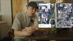 Person gesturing toward aerial satellite map displayed on tablet screen.