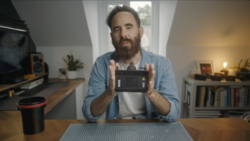 Bearded man holding a camera controller while seated at a desk in a home studio.