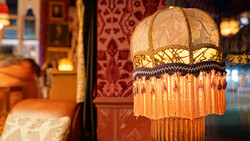 Ornate vintage pendant lamp with cream fabric shade, gold embroidered trim, and long orange beaded fringe hanging in warm interior space.