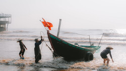 Fishermen in water pulling ropes attached to a green and black wooden boat with an orange flag in rough seas.