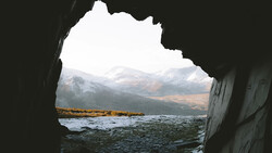 Snow-capped mountains and a rocky coastline viewed through a natural cave opening.