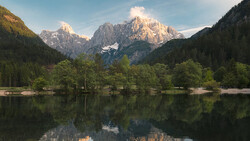 Alpine lake with forested shoreline and snow-capped mountain peaks reflected in still water.