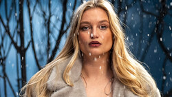 Woman with long blonde hair in falling snow against blue winter backdrop.