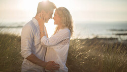 Couple embracing during golden hour on a beach with tall grass and ocean backdrop.